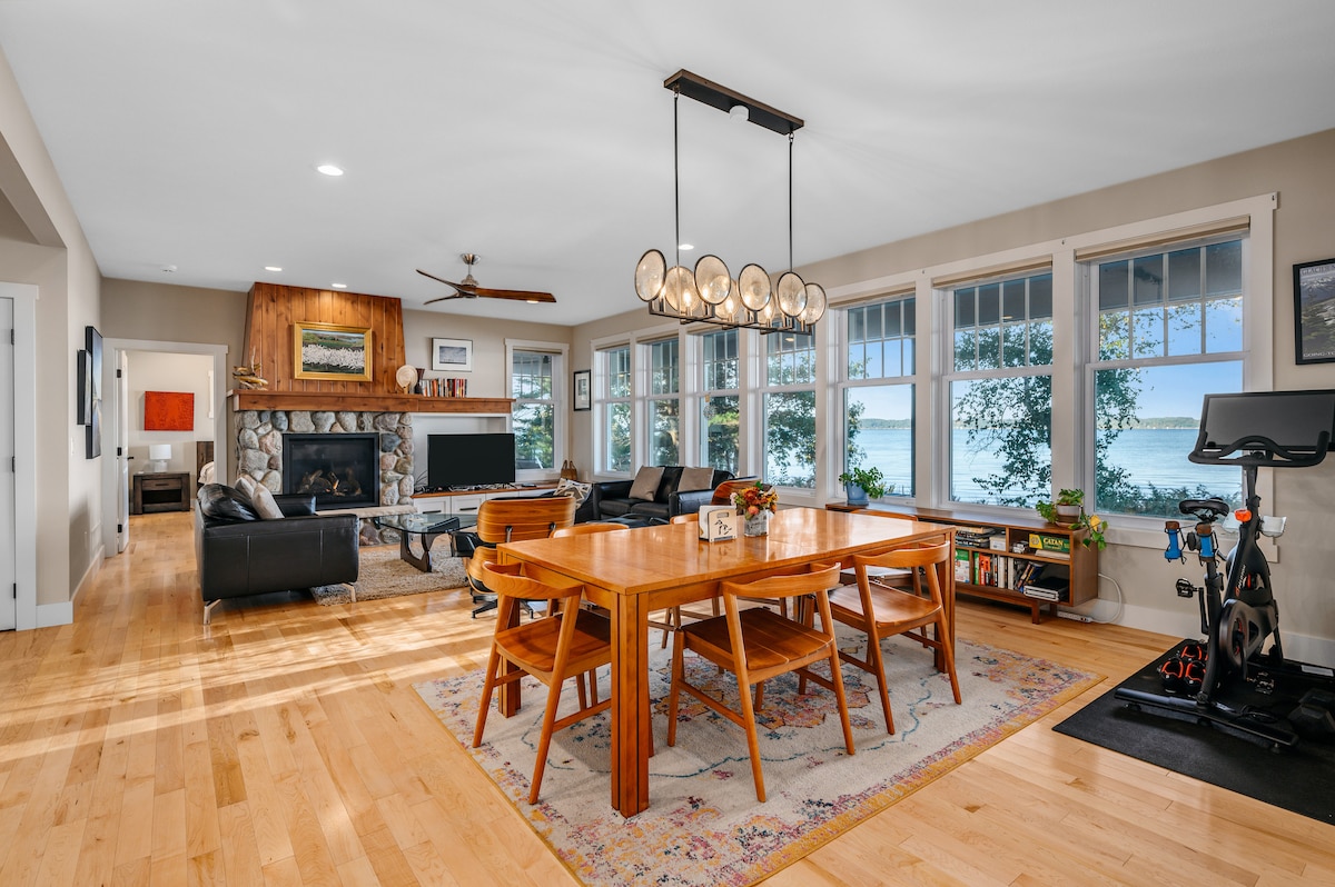 A modern and open living area is highlighted by large windows overlooking the water, allowing natural light to fill the space. The dining table with wooden finishes is surrounded by comfortable chairs, while a cozy seating arrangement with dark furniture is positioned near a stone fireplace.