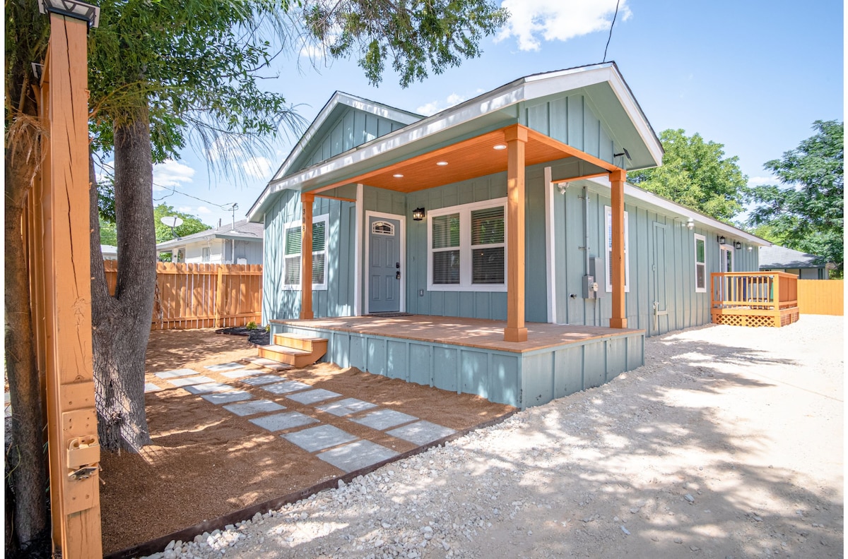 A welcoming exterior view of a single-story home features a covered porch with wooden beams. Light blue siding complements the white trim. A stone pathway leads to the entrance, surrounded by a gravel yard, trees, and privacy fencing in the background.