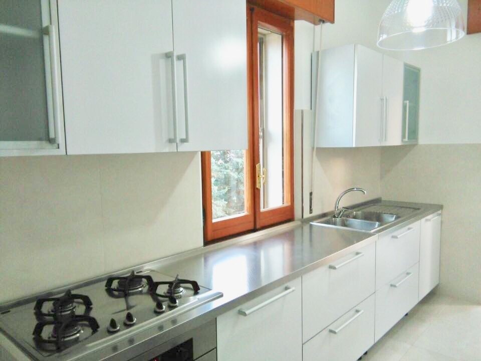 A modern kitchen features sleek white cabinetry and a stainless steel countertop. A gas stove and sink are integrated into the design, while a large window allows natural light to fill the space, creating a bright and welcoming environment.
