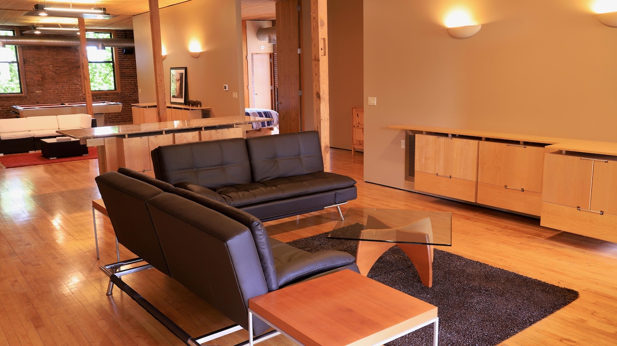 The living area features a modern design with sleek black leather seating arranged around a minimalist glass coffee table. Wooden cabinetry provides storage, while natural light brightens the space through large windows. A layered rug adds texture to the wooden flooring.