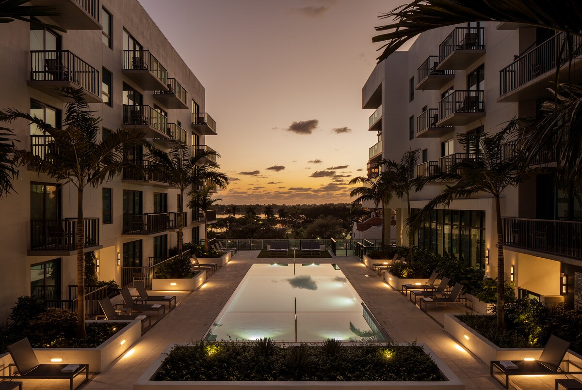 An inviting pool area is framed by modern buildings, complemented by palm trees and soft landscaping. Subtle lighting enhances the tranquil ambiance, while the horizon showcases a blend of warm sunset hues over a distant view of water.