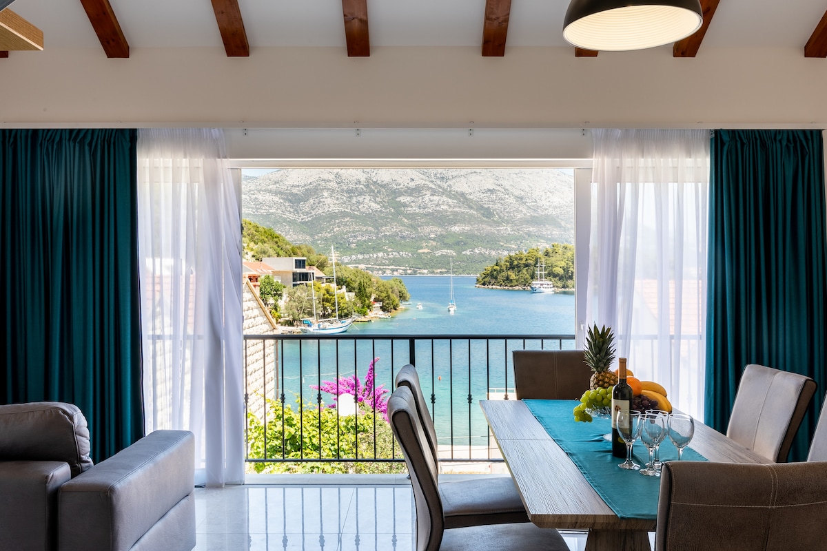 A spacious living area showcases a dining table set for guests, with vibrant blue-green curtains framing a stunning view of the water and nearby coastline. Natural light enhances the space, creating a pleasant atmosphere for dining or relaxation.