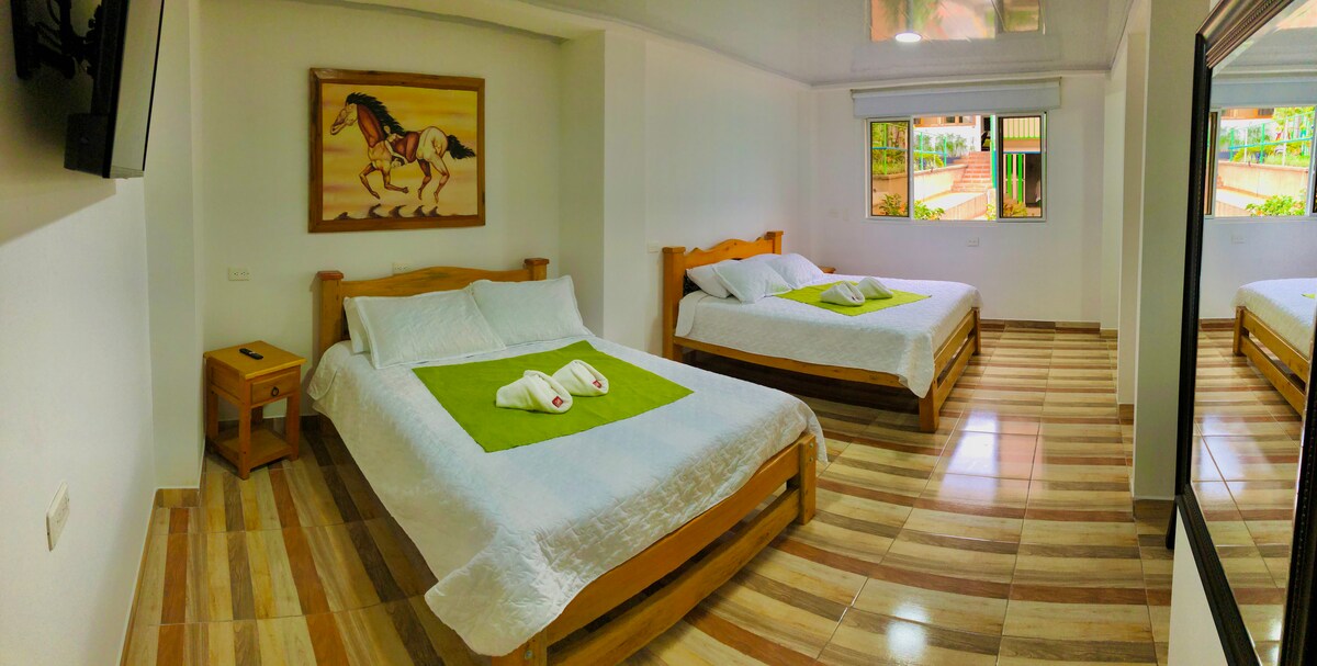 The spacious room includes two large beds with white linens and green accents. A wooden nightstand is positioned beside one bed, and a flat-screen TV is mounted on the wall. Large windows offer natural light and views of the outside.