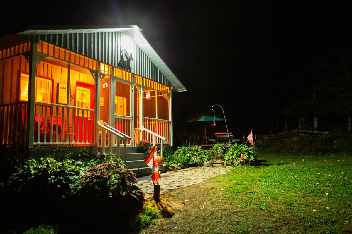 The cottage's inviting exterior is illuminated at night, showcasing a covered porch adorned with vibrant rocking chairs. Flanking the entrance are Canadian flags, while the landscaped yard features plants and a stone pathway leading to a fire pit area surrounded by greenery.