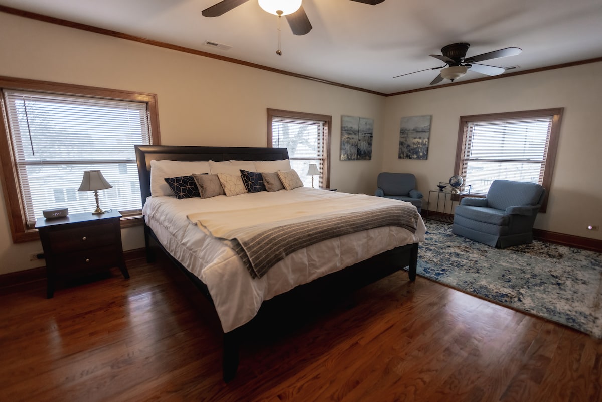 The spacious master bedroom features a large bed adorned with decorative pillows and a soft throw blanket. Two windows allow natural light to fill the room, and a cozy seating area includes a chair and a side table. A textured area rug adds warmth to the hardwood floor.