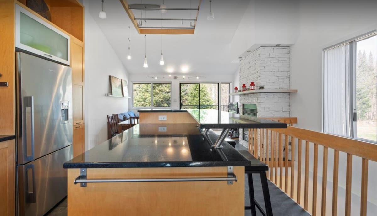 A modern kitchen features a sleek dark countertop and stainless steel appliances. Large windows provide natural light and views of the surrounding greenery. The open layout connects the kitchen to the living area, where a stone fireplace is visible in the background.