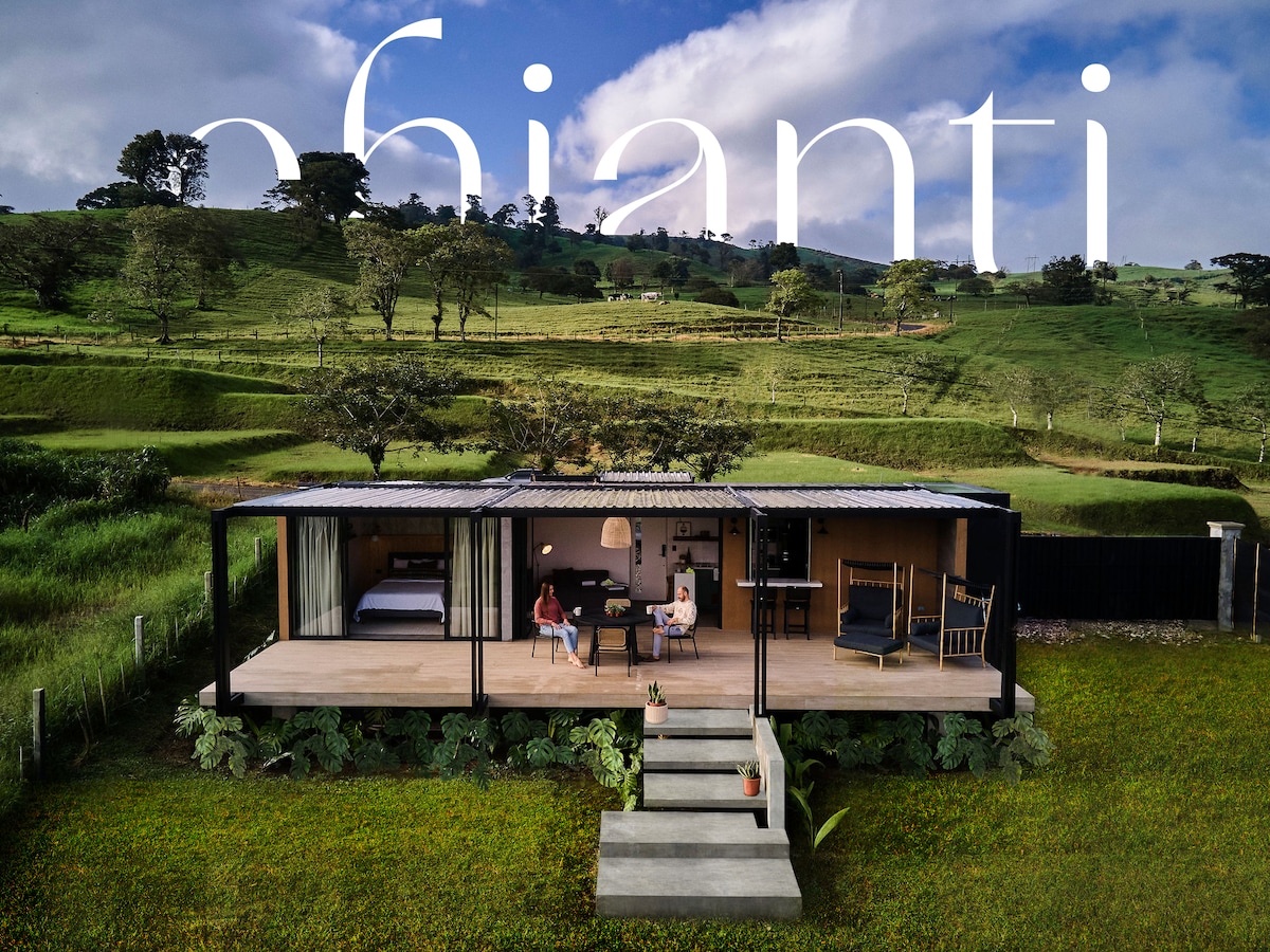 A contemporary mountain home features large glass openings, allowing natural light to fill the living spaces. The exterior is surrounded by lush greenery and scenic hills. A few outdoor steps lead to a welcoming entrance, creating a connection with the surrounding landscape.