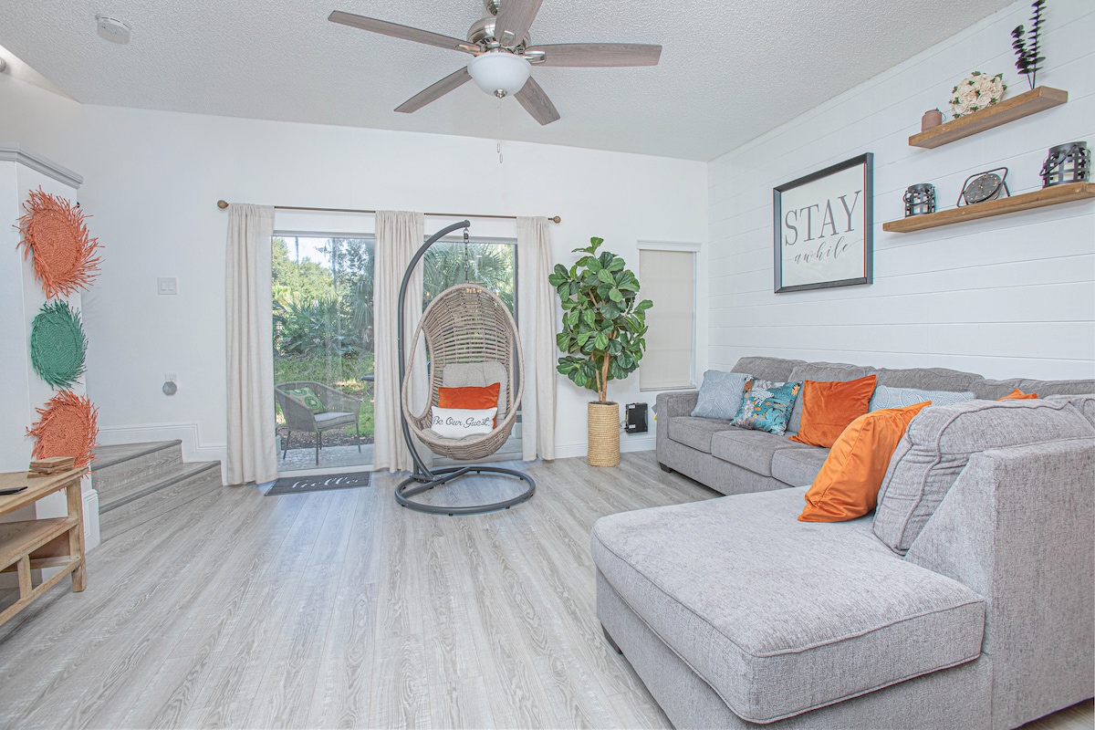 A spacious living area features a large sectional sofa adorned with bright orange and teal accent pillows. A hanging chair adds a unique touch, while natural light pours in through sliding glass doors leading to an outdoor space. Potted plants bring a refreshing vibe.