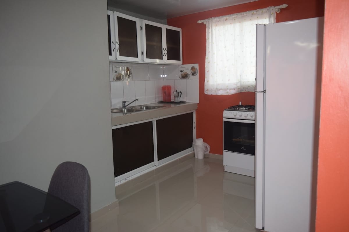 A compact kitchen is displayed, featuring white cabinets and a stainless-steel sink. A gas stove is positioned next to a modern refrigerator. Natural light enters through a curtained window, illuminating the polished tiled floor.