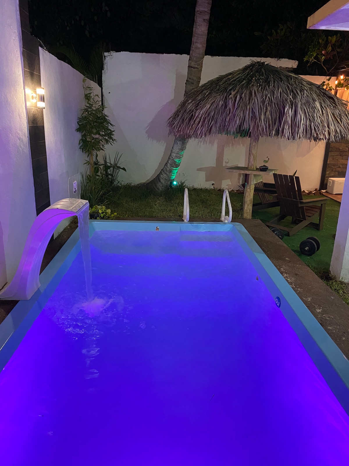 A cozy outdoor pool area features a softly illuminated pool, highlighted in calming purple hues. A thatched-roof shade structure provides a tranquil space, while a small seating area with wooden chairs is positioned nearby. Lush greenery surrounds the setting, enhancing the serene atmosphere.
