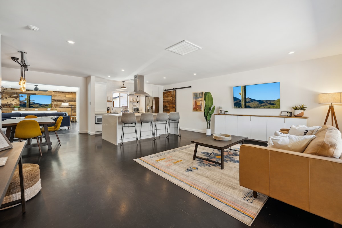 An open floor plan is showcased, featuring a modern kitchen with a large island and seating for four. Natural light fills the space through large windows, illuminating the stylish living area with comfortable seating and vivid artwork. A dining area is visible in the background.