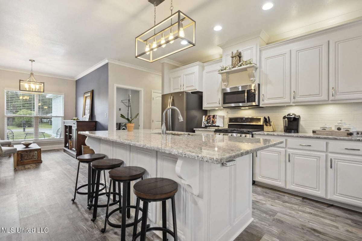 The kitchen features white cabinetry and a large island with seating for four. Stainless steel appliances are integrated into the design, while a modern light fixture illuminates the space. Natural light enters through a nearby window, enhancing the inviting atmosphere.