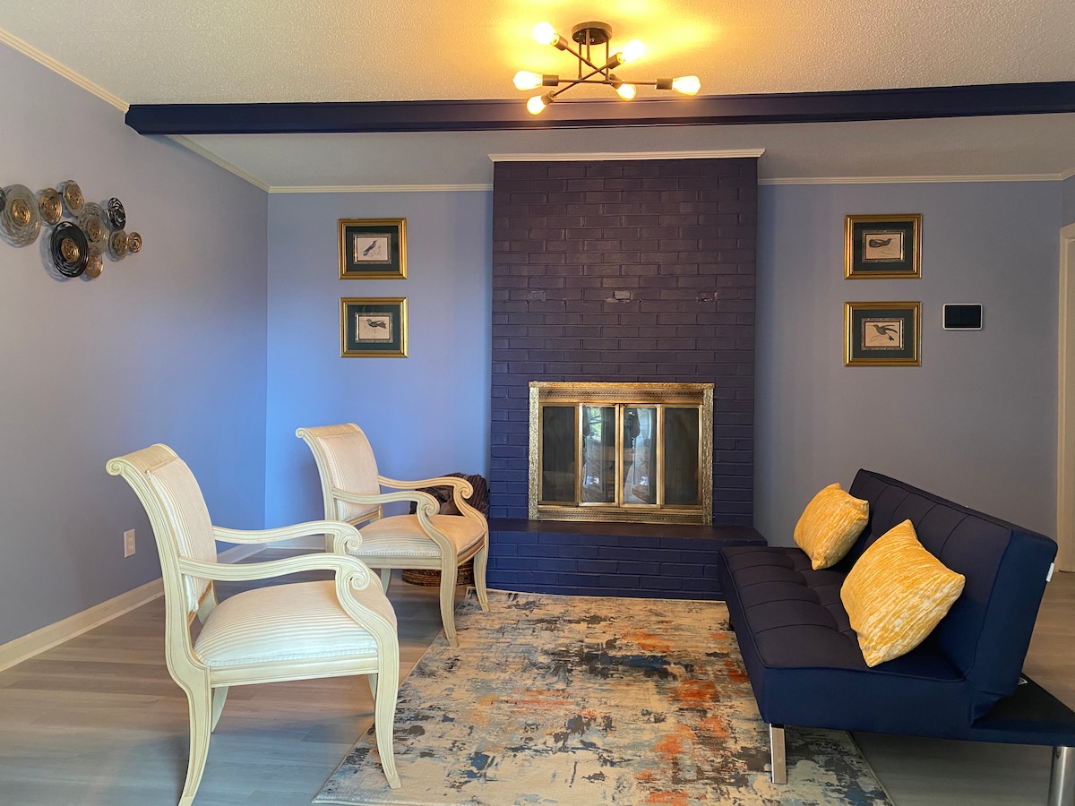 A comfortable living area features a stylish navy sofa accompanied by two elegant armchairs. The blue walls are complemented by decorative wall art and framed pictures. A modern fireplace with a gold frame adds a focal point, while a colorful area rug completes the inviting space.
