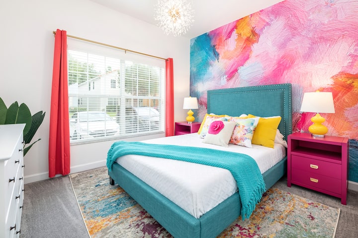 Dormitorio vibrante y colorido con una pared decorativa audaz, una cama tapizada de color verde azulado y una decoración alegre. Los toques brillantes de rosa, amarillo y turquesa crean un ambiente acogedor y juguetón, perfecto para una estancia alegre.