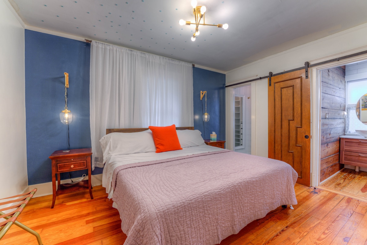 A spacious bedroom features a large bed with a light purple quilt and an orange accent pillow. Soft lighting is provided by pendant lights on either side. A wooden nightstand and a partially visible closet add to the room's functionality. The walls are painted in calming blue, complemented by natural light filtering through sheer curtains.