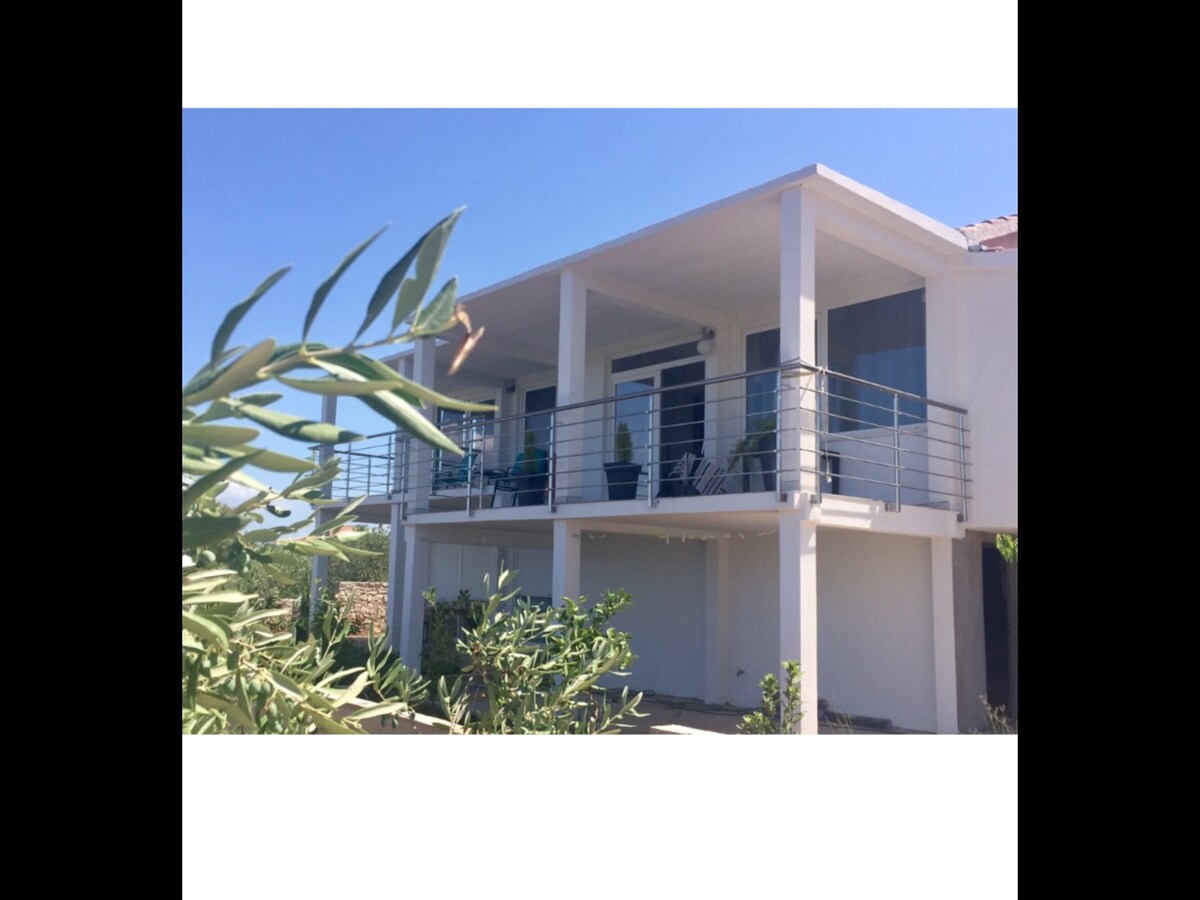 The exterior of a modern two-story house is shown, featuring a spacious balcony with railing. Green plants frame the image, and comfortable seating is visible on the balcony. The sky is clear, providing natural light to the structure.
