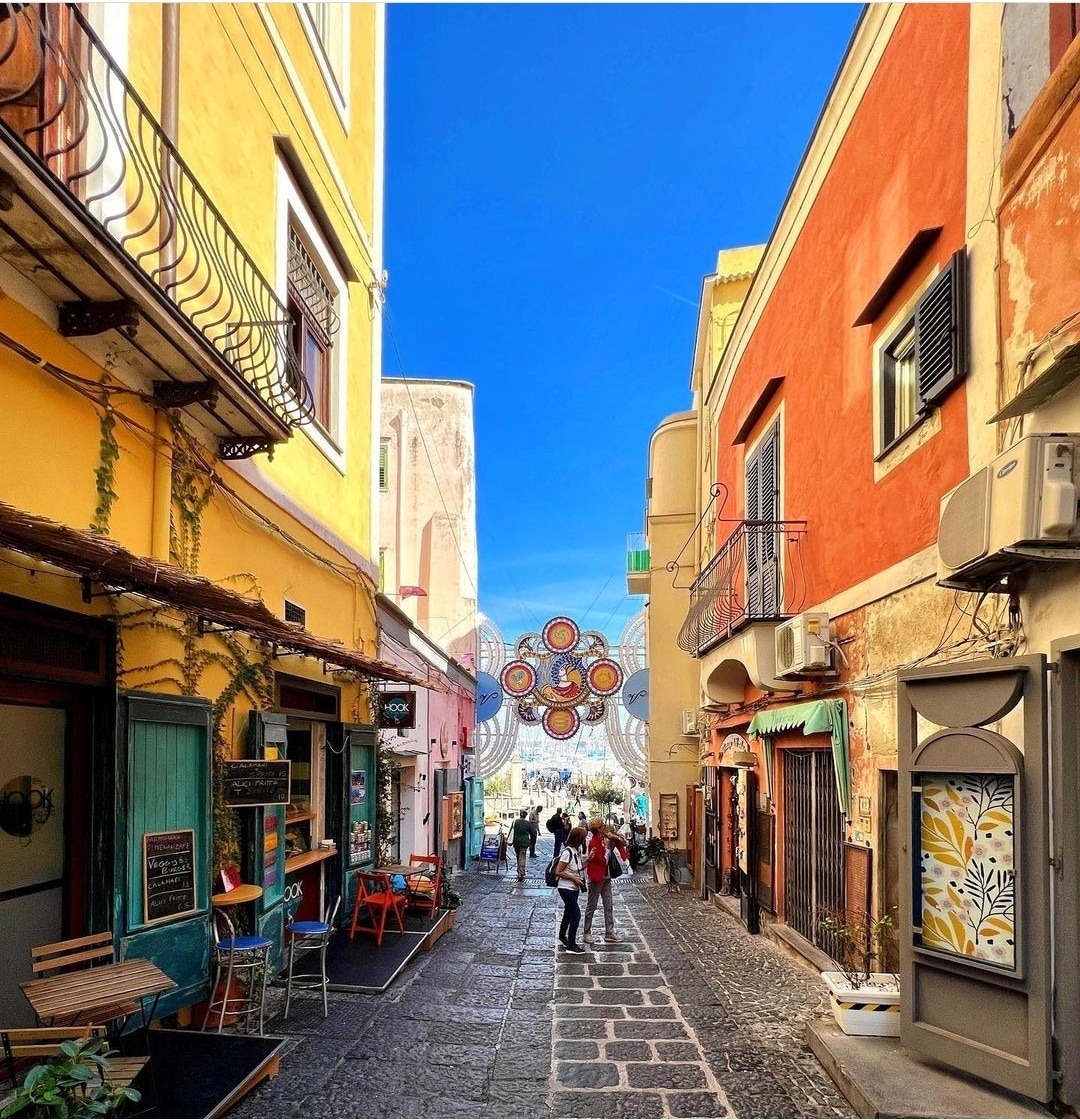 A vibrant alleyway is flanked by colorful buildings in shades of yellow and orange, with decorative elements and greenery present. A cobblestone path leads through the area, where visitors can be seen strolling and admiring the local shops and festive decorations overhead.