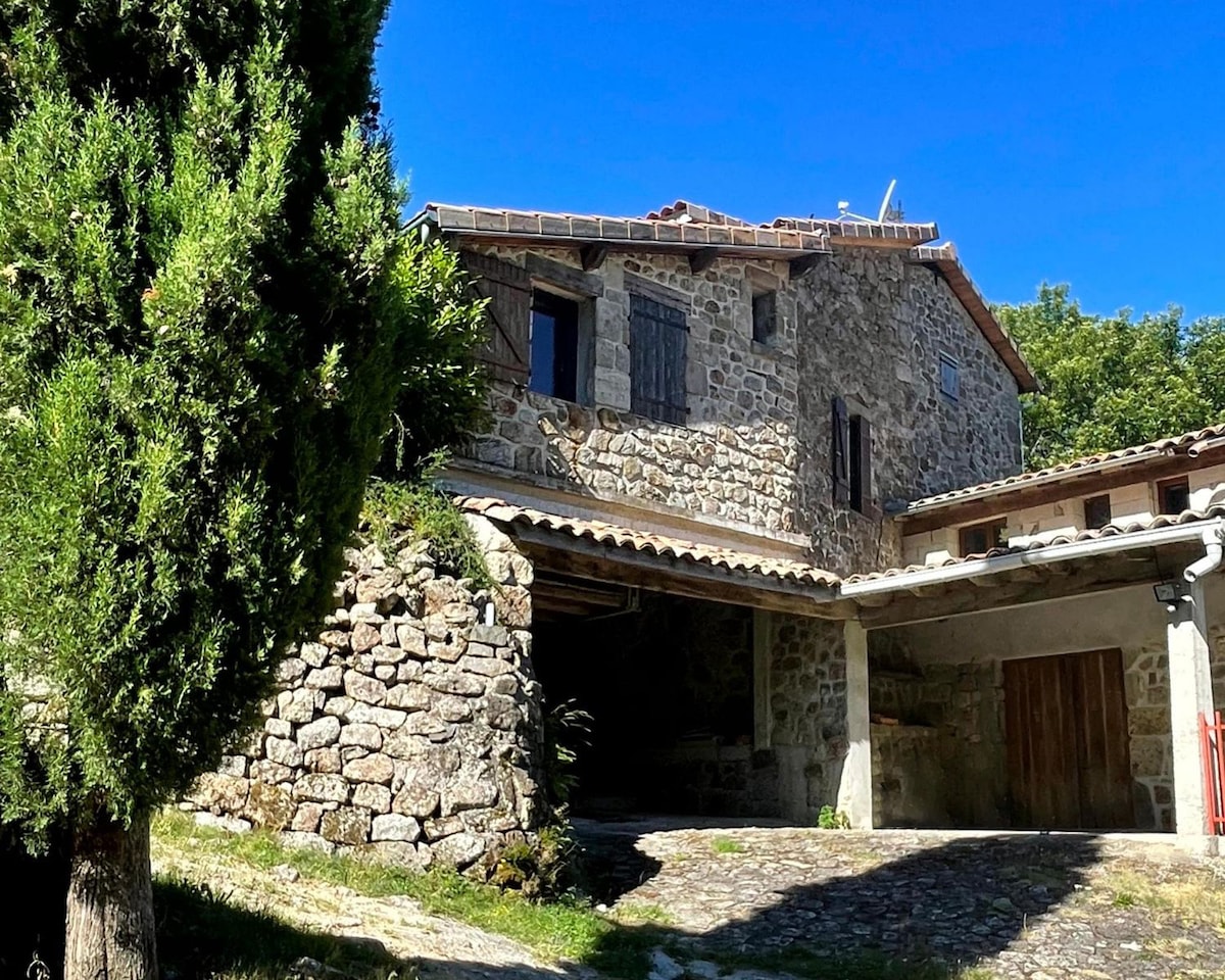 A charming stone house sits nestled among greenery, with a weathered exterior and wooden shutters. A sloped, grassy area leads up to the structure, complemented by a gravel path and a tree providing shade. Clear blue skies are visible overhead.