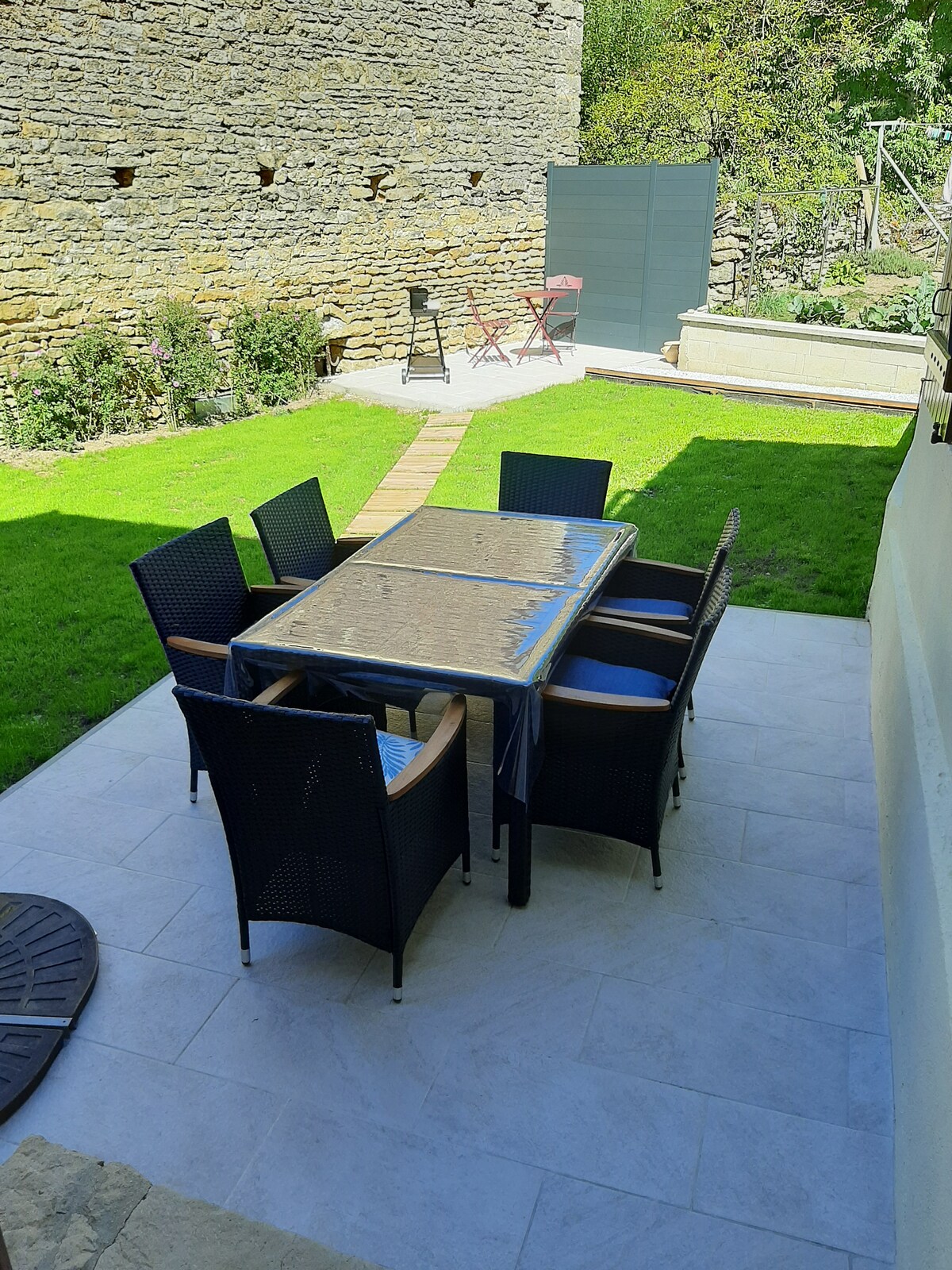 An outdoor dining area is arranged with a rectangular glass-top table surrounded by six wicker chairs on a tiled patio. Lush green grass stretches across the yard, complemented by low stone walls and blooming plants in the background.