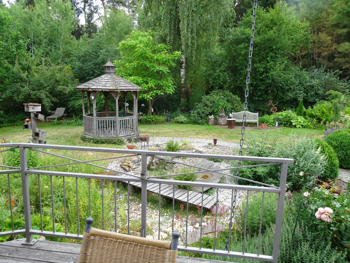 A well-maintained garden is shown, featuring a central gazebo surrounded by lush greenery. Comfortable seating is visible, with stone paths leading through flowerbeds. Various trees and shrubs create a natural backdrop, providing a serene atmosphere for relaxation.