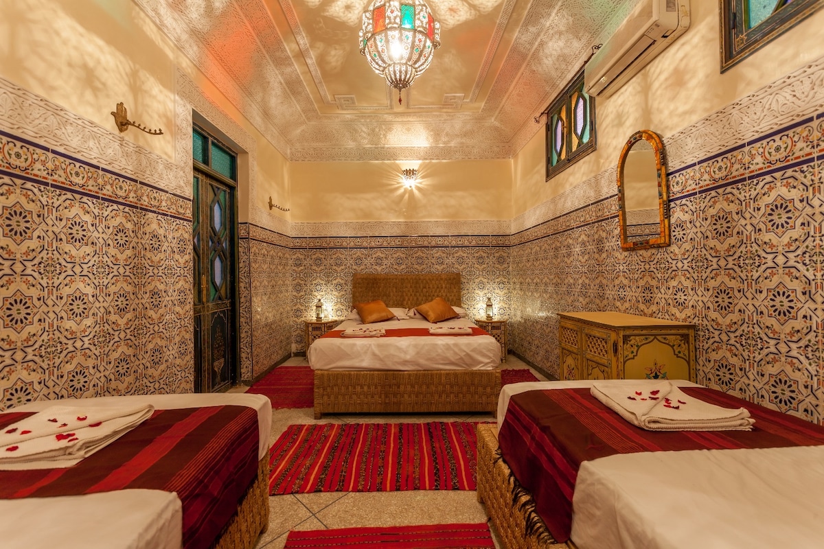 A family room features a spacious double bed and two single beds, surrounded by intricately patterned tiled walls. Soft lighting highlights the room's vibrant decor, with traditional textiles adorning the floor. A mirror and wooden dresser add functional elegance, creating a cozy atmosphere.