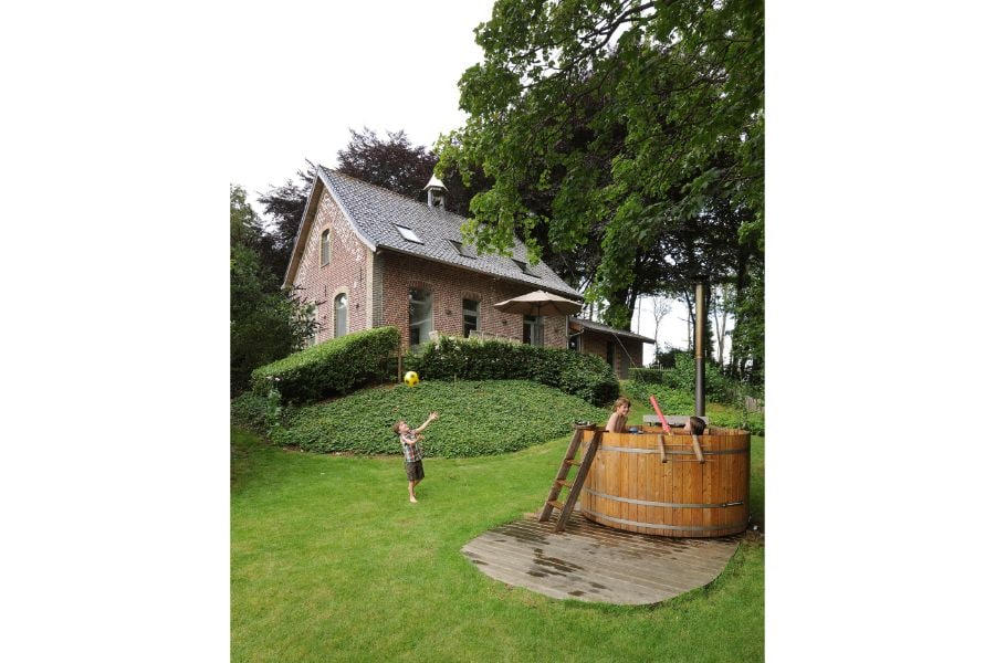 A spacious garden surrounds a charming brick house, featuring green hedges and trees. A wooden hot tub is positioned nearby, accessible by a small wooden ladder. Two children play joyfully on the grass, while a patio umbrella provides shade beside the house.