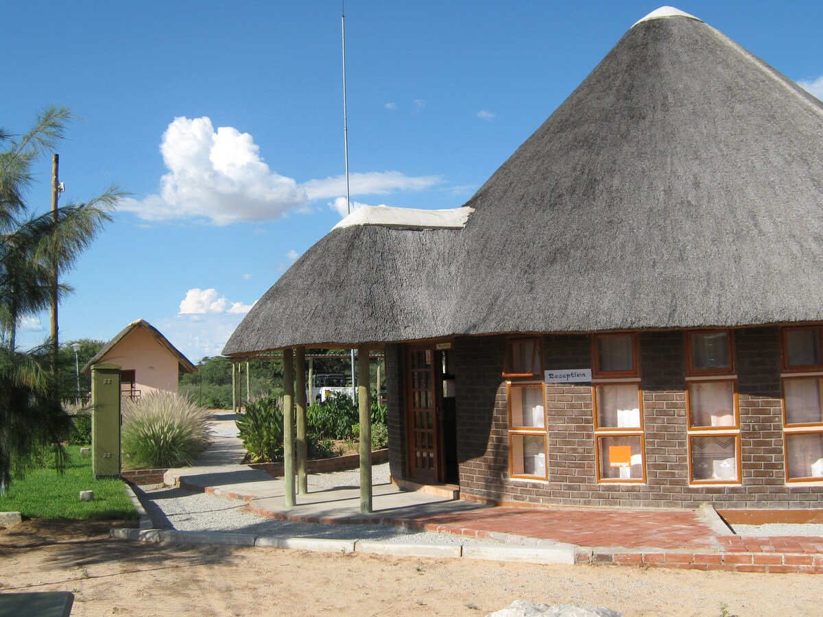 A thatched-roof structure is prominently featured, showcasing a combination of brick and wooden accents. Large windows allow natural light to fill the interior. Surrounding greenery and a pathway lead to additional buildings in the background, enhancing the inviting outdoor space.