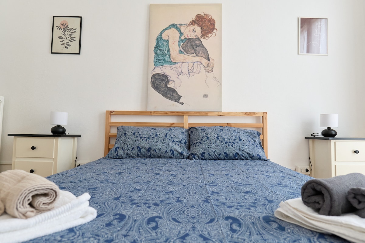 The bedroom features a spacious bed with a decorative blue coverlet and two pillows. Nightstands with lamps are positioned on either side. A piece of wall art is displayed above the bed, complementing the room's neutral walls and ambiance.