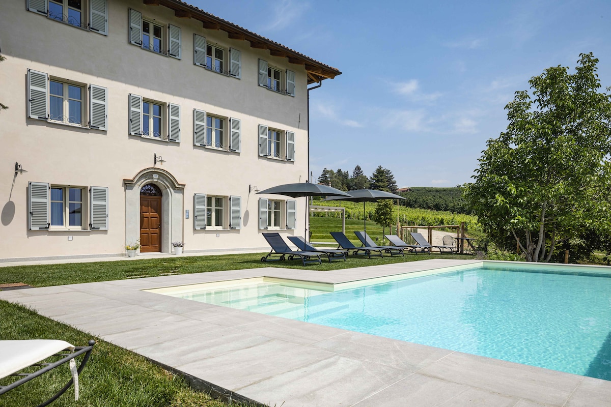 A charming villa is depicted with a tranquil pool surrounded by sun loungers under umbrellas. The light-colored exterior features multiple windows with shutters, and a welcoming entrance leads the eye toward lush greenery in the background.