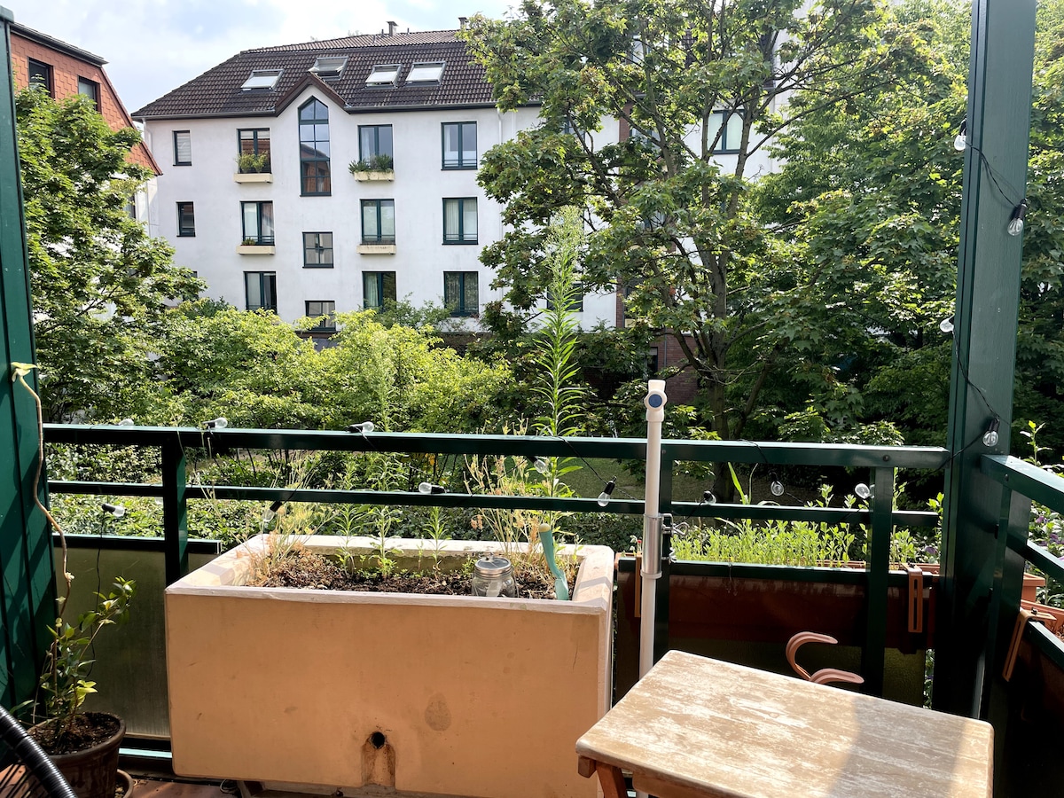 A balcony showcases a view of lush greenery and surrounding buildings. A planter box is visible, featuring various plants, while a small table sits nearby, providing a spot to enjoy the serene environment. String lights add a subtle touch to the space.