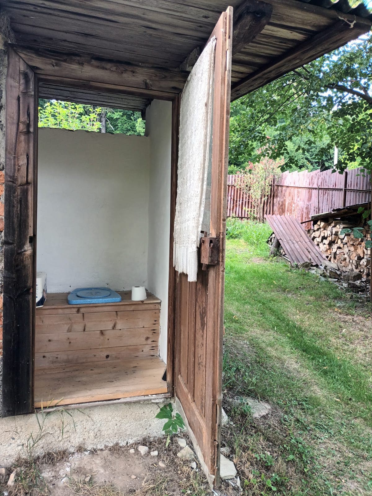 An outdoor toilet is shown, featuring wooden walls with a rustic door that is partially opened. Inside, a blue toilet seat is positioned on a wooden platform. The entrance displays a simple curtain for privacy, while the surrounding greenery and a wooden fence create a natural setting.