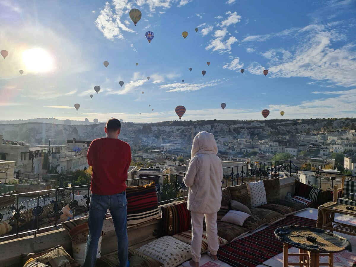 An expansive terrace is furnished with comfortable seating, offering an unobstructed view of colorful hot air balloons soaring in the clear sky over Cappadocia. Soft textiles and traditional patterns adorn the seating area, enhancing the scenic experience. The landscape features rolling hills and quaint buildings in the background.