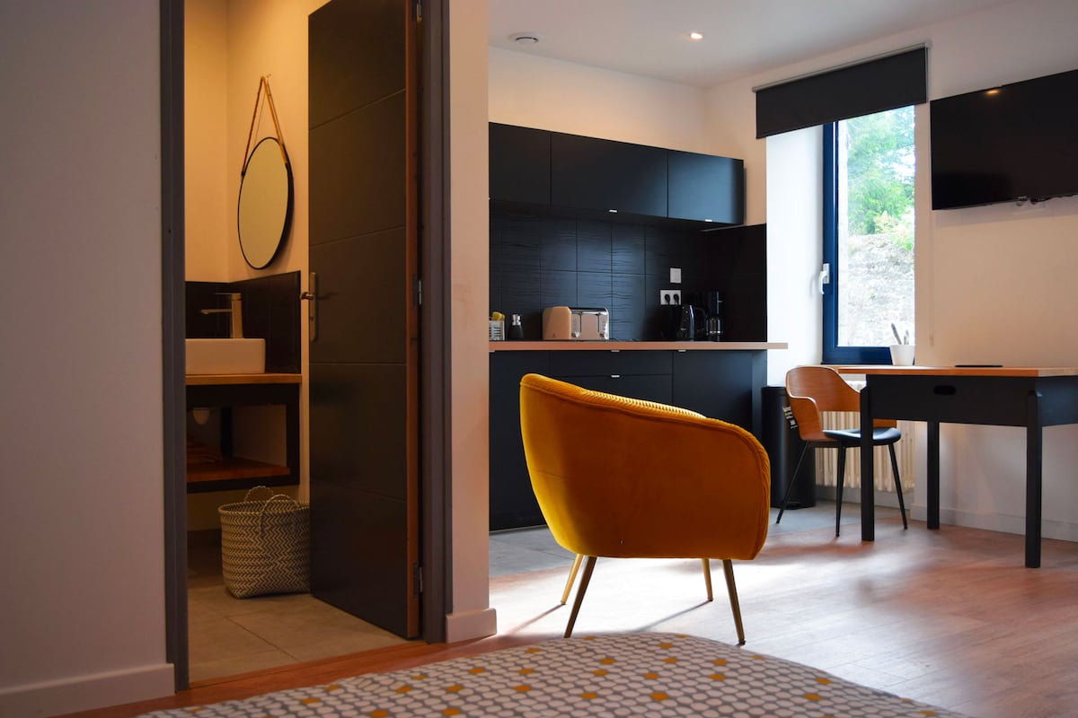 A modern studio is displayed, featuring a cozy seating area with a bright yellow armchair facing a minimalist kitchen equipped with essential appliances. A wooden table is positioned nearby, and a doorway leads to a well-appointed bathroom that includes a mirror. Natural light streams in through a large window.