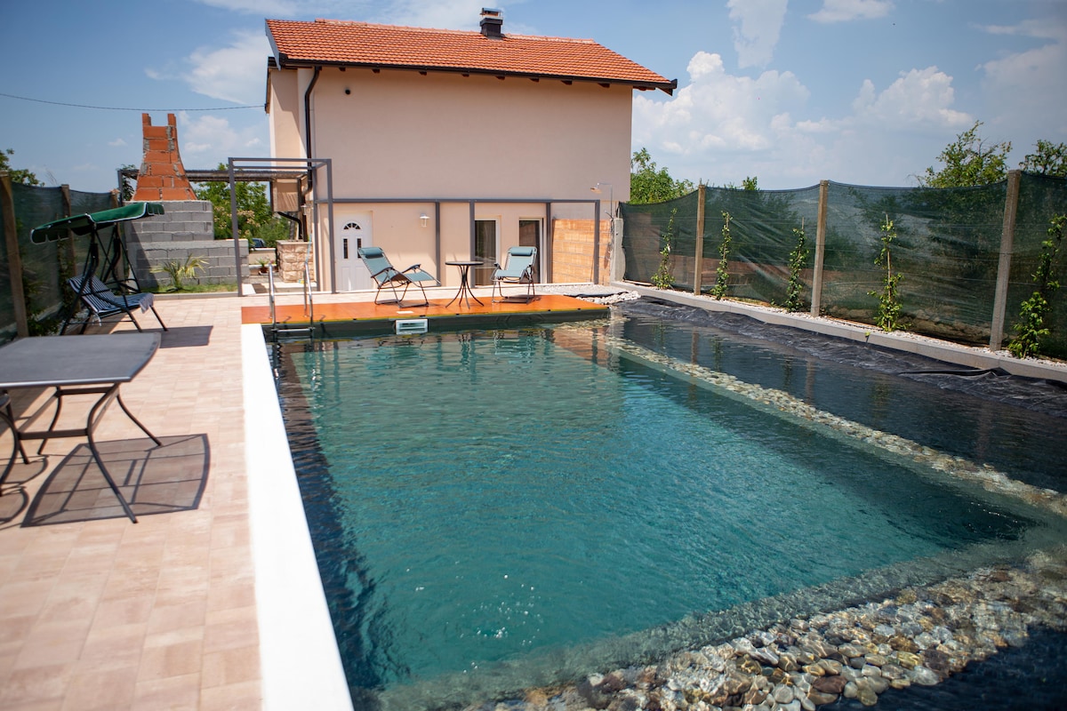 A spacious private pool is surrounded by a sunbathing area with chairs and a table. The pool features a stone-bordered edge and is encased by a green fence, providing a sense of privacy. A modern holiday home is visible in the background.