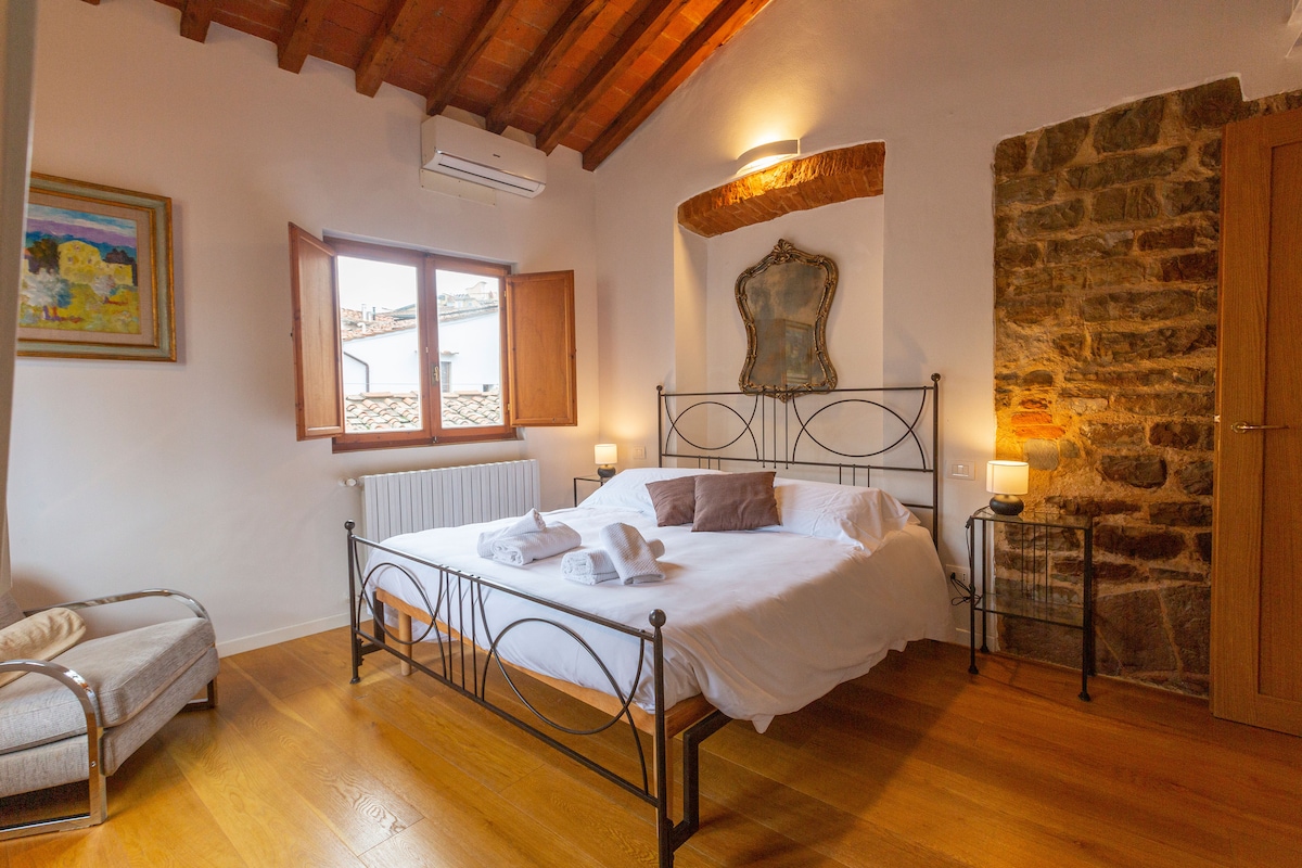 A spacious bedroom features a wrought-iron bed dressed in white linens, flanked by bedside lamps. Natural light enters through wooden window shutters, highlighting the stone wall to the right. A painting adds a touch of color, while a cozy armchair sits in the corner.
