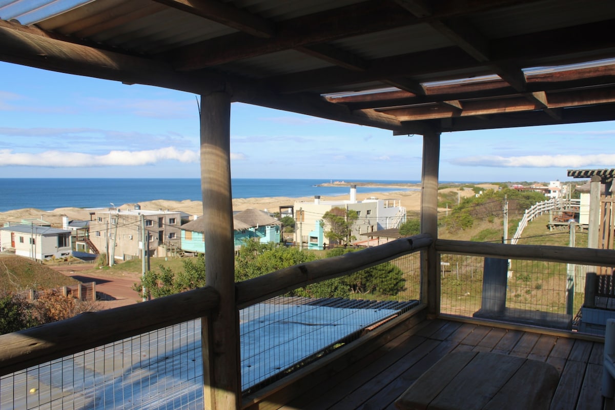 A wooden balcony offers a view of the coastline and sandy beach. The ocean stretches beyond, framed by a line of buildings along the shore. Gentle waves are visible, with the sky featuring soft clouds and a hint of blue.