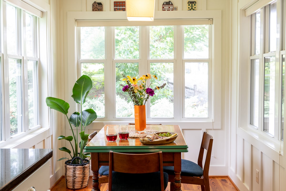 A cozy dining area is illuminated by natural light streaming through large windows. A wooden table is set with glasses and snacks, accompanied by a vase of fresh flowers. A potted plant adds a touch of greenery to the space.