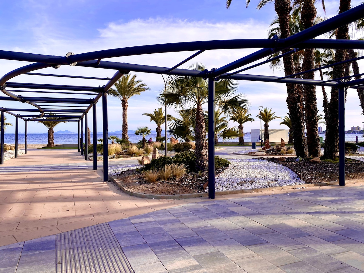 A pedestrian walkway adorned with palm trees leads towards the shoreline. The area features modern architectural elements, with a structured overhead canopy and landscaped gardens. The calm waters of the sea are visible in the background, creating a serene coastal atmosphere.