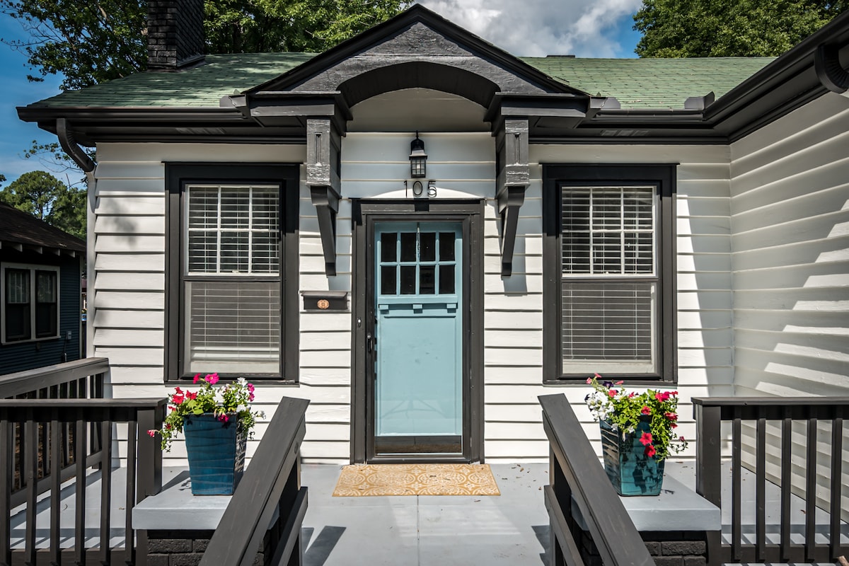 The front entrance of the bungalow is framed by two flower pots containing colorful blooms. A turquoise door, adorned with a lantern, welcomes guests. The entrance features a small mat on a covered porch with a simple railing, accentuating the welcoming appearance of the home.