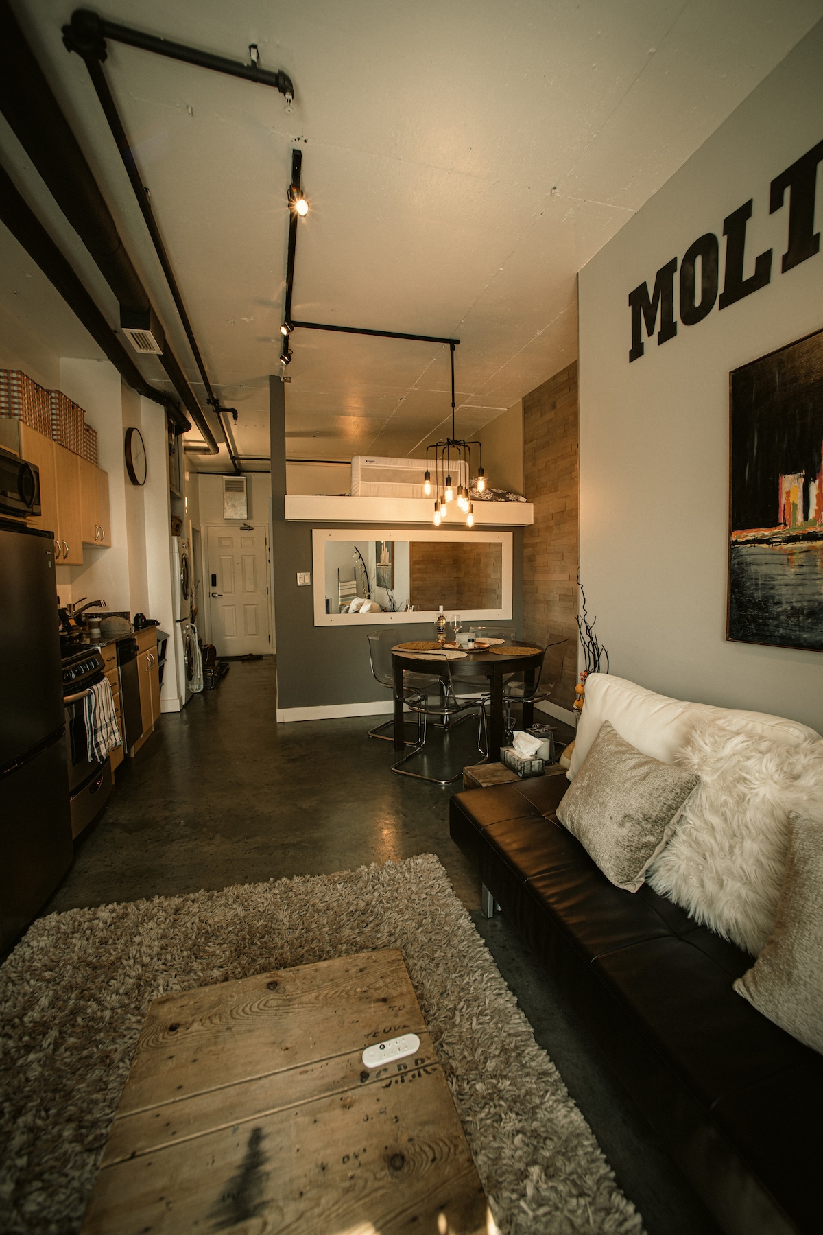 The living area features a cozy black couch adorned with decorative cushions, set against a backdrop of industrial-style walls. A rustic wooden coffee table sits on a plush area rug, while a chic dining table with metal accents and modern light fixtures provides a functional and stylish space.