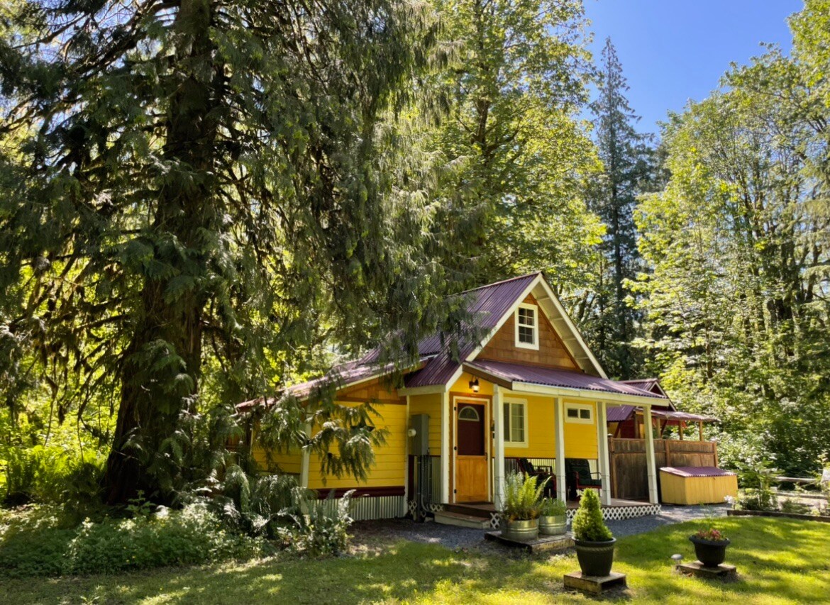 A cheerful cabin surrounded by lush green trees showcases a vibrant yellow exterior. Sunlight filters through the foliage, creating a warm atmosphere. The porch area is partially visible, inviting relaxation in a tranquil setting, while the surrounding garden features a potted plant.