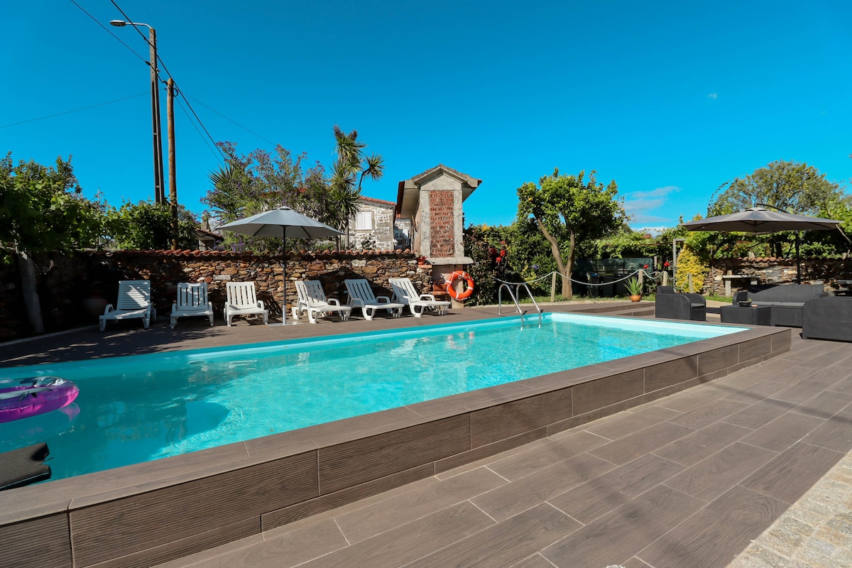 An outdoor space features a refreshing pool surrounded by sun loungers and shaded areas under umbrellas. Lush greenery and trees create a serene atmosphere. A wooden deck frames the pool, enhancing the outdoor leisure area with seating arrangements, inviting relaxation and enjoyment.