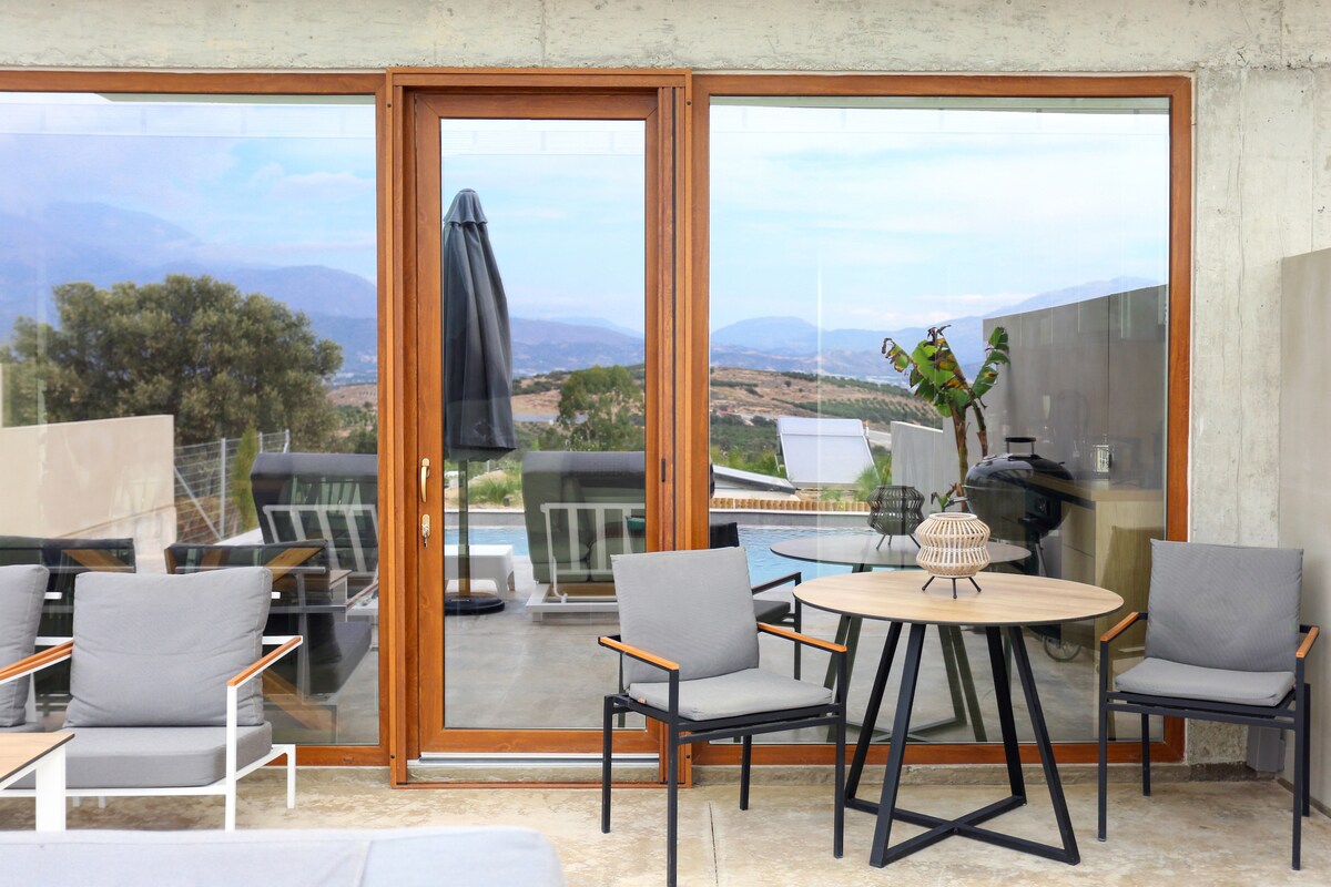 A patio area features a round wooden table surrounded by four grey armchairs. Large glass doors provide a view of the private swimming pool and surrounding landscape, while a black umbrella stands ready for shade.