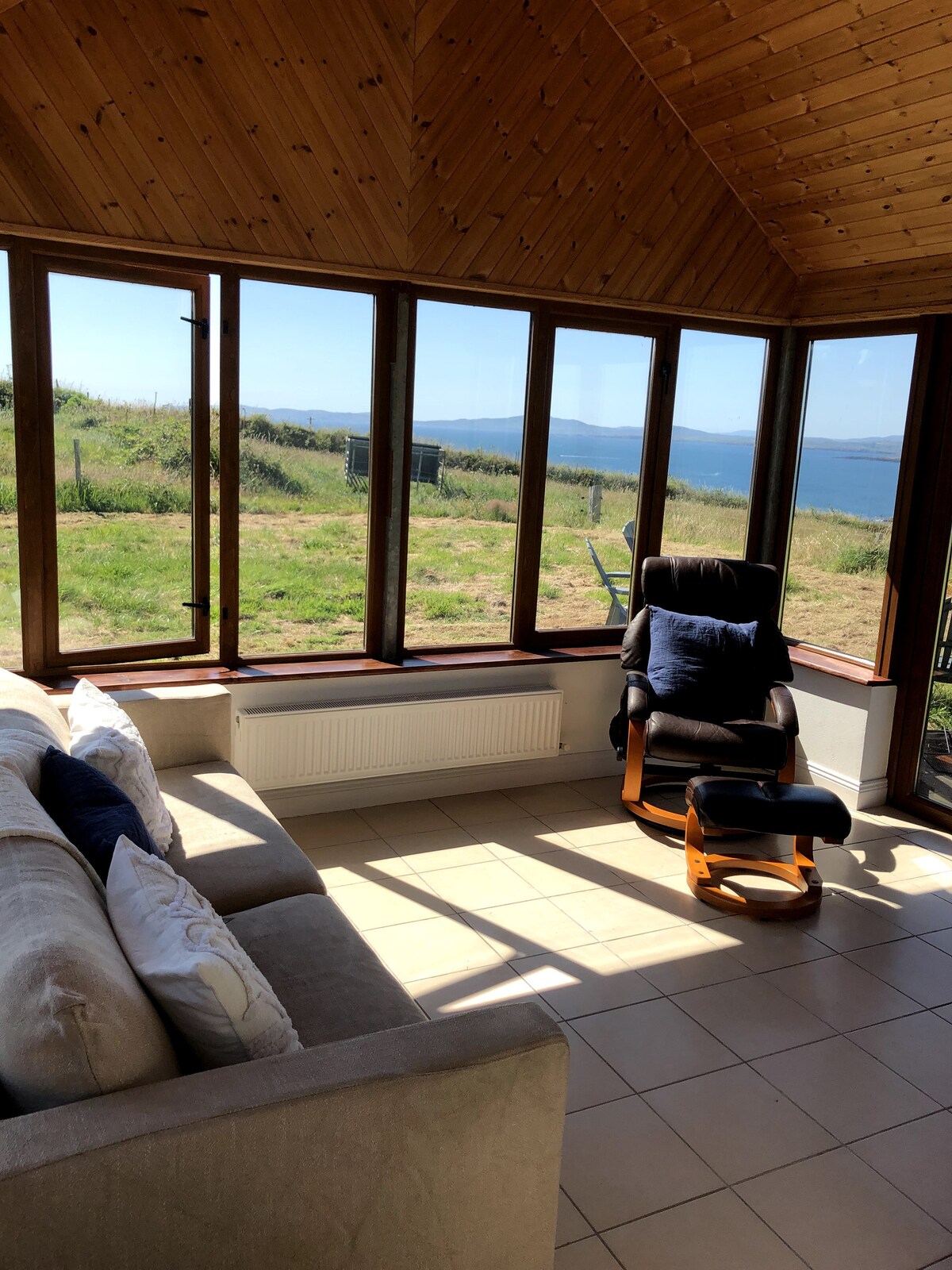 A sunlit sunroom features large glass windows that offer sweeping views of the Atlantic Ocean. A comfortable sofa and a rocking chair are positioned on the tiled floor, creating a cozy spot for relaxation while enjoying the surrounding landscape.
