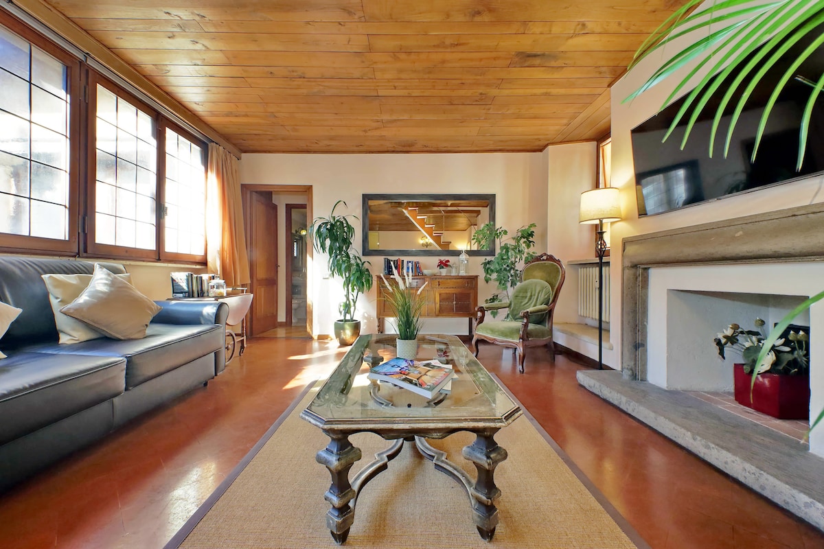 A spacious living area features a wooden ceiling and large windows allowing natural light to fill the room. A comfortable dark sofa is paired with a vintage coffee table, while potted plants add a touch of greenery. A smart TV is mounted on the wall for entertainment.