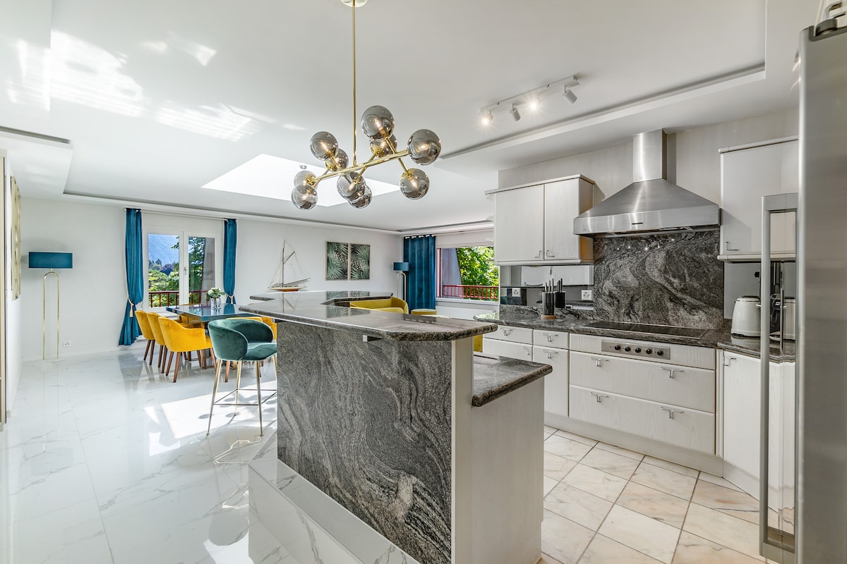 A modern kitchen and dining area are showcased, featuring a marble island and sleek cabinetry. A spacious dining table is surrounded by colorful chairs, and natural light floods the space through large windows. Bright blue curtains frame the windows, enhancing the airy ambiance.