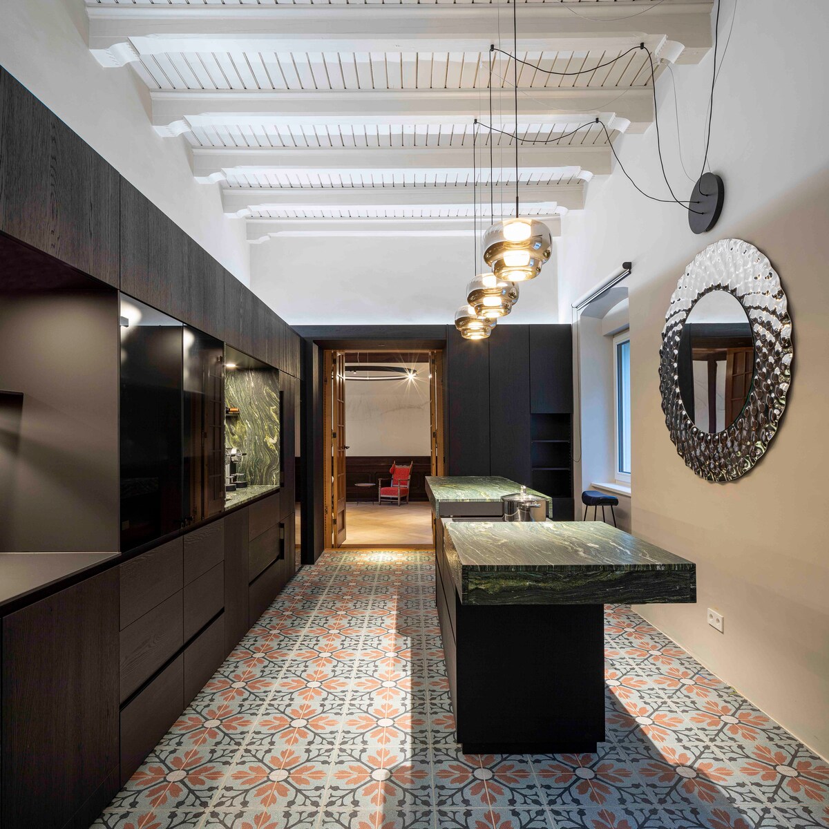 The elegant kitchen features a large central island topped with dark stone, surrounded by sleek cabinetry. A patterned tile floor adds texture, while a round mirror reflects the space. Pendant lights hang from a high ceiling, enhancing the modern design.