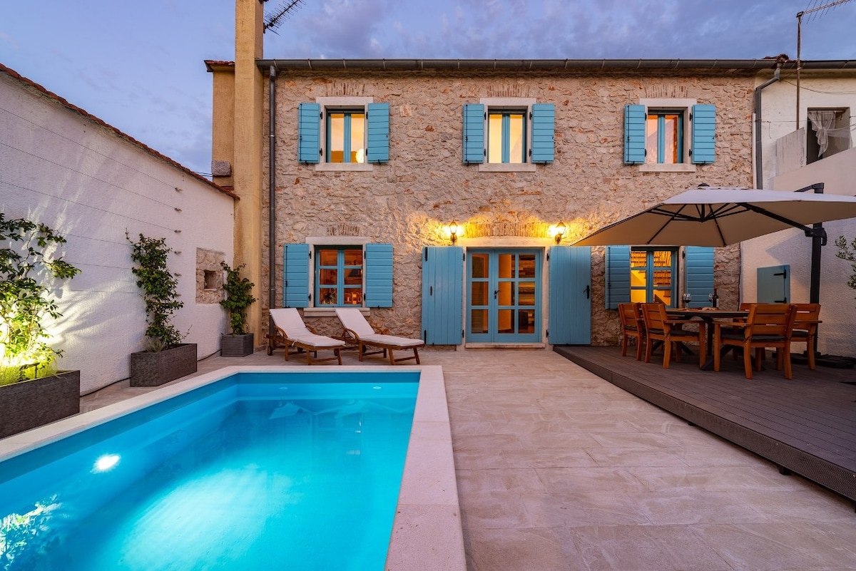 A traditional stone villa exterior is highlighted by blue shutters and warm lighting. The inviting pool is surrounded by deck chairs, while a dining area with wooden furniture provides a comfortable space for outdoor meals. A shaded umbrella adds to the outdoor relaxation atmosphere.