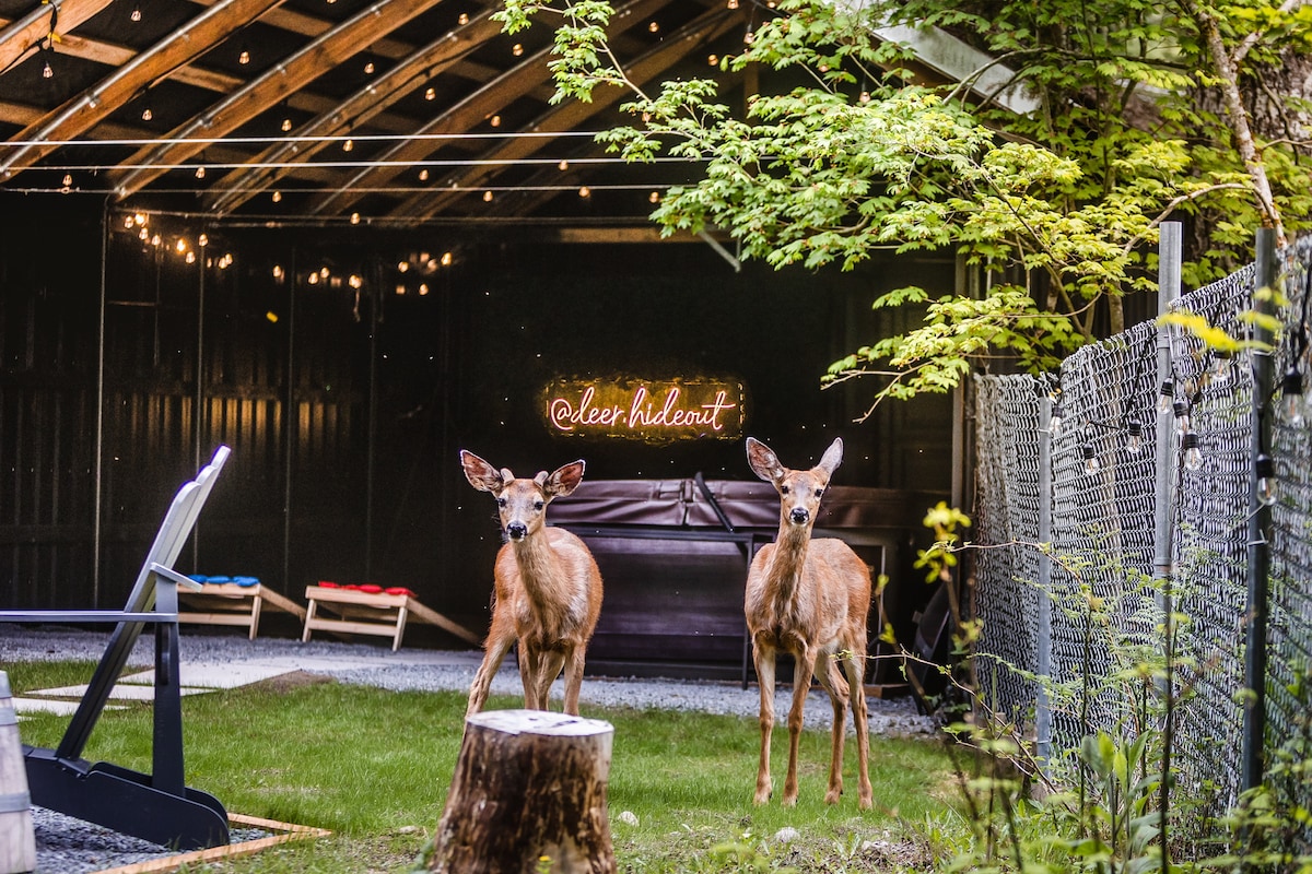 Two deer are seen standing near a wooden hot tub, with festive string lights illuminating the background. A cozy seating area with lounge chairs and a tree stump adds to the inviting outdoor ambiance, framed by lush greenery.