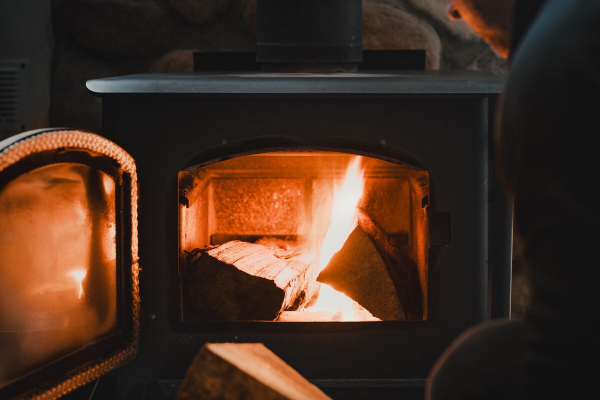 A wood-burning stove is shown, featuring flames illuminating the inside with warmth. Logs are stacked inside, and the gray stone backdrop adds a rustic touch. The stove's glass door allows for the flickering firelight to be observed, creating a cozy ambiance.